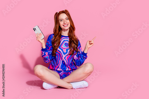 Full length photo of lovely young lady sit floor hold gadget point empty space dressed blue print garment isolated on pink color background photo