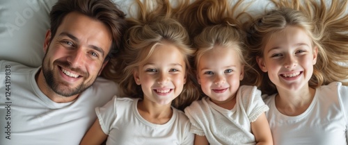 A cheerful family of four lies together on a soft, white bed, their hair cascading around them like golden waves. The radiant smiles of the father and his three daughters create a heartwarming scene