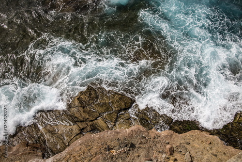 Aerial view of waves crashing against rocks on a shoreline. Dynamic patterns and textures in the water and rocks.