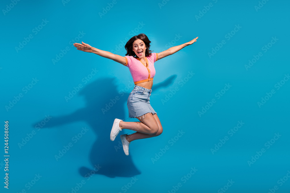 Excited young woman jumping with joy in a stylish casual outfit against a vibrant blue background Excited young woman jumping with joy in a stylish casual outfit against a vibrant blue background