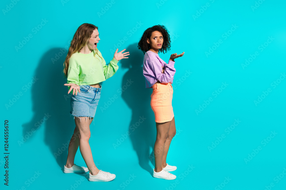 Obraz premium Two young women gesturing expressively against a turquoise background, showcasing diverse styles and trends in casual fashion.