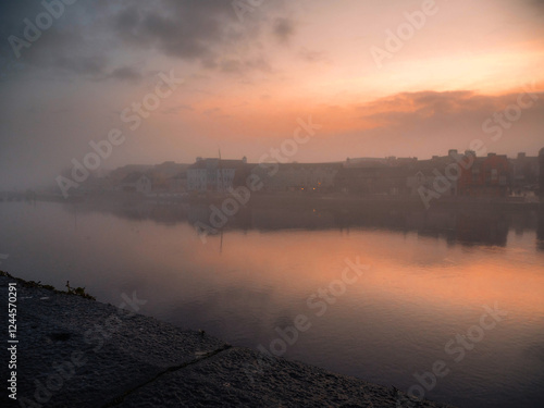 Athlone city riverside at warm dusk. Cloudy sky reflects in river Shannon. Popular old town, shopping, educational and industrial center. Tourist and travel hub. Fog over river creates surreal look photo