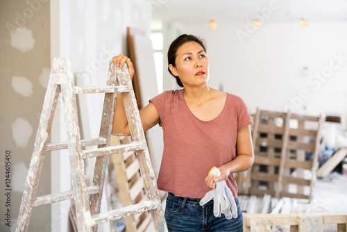 Obraz na płótnie Thoughtful asian woman standing at stepladder in apartment during repair works