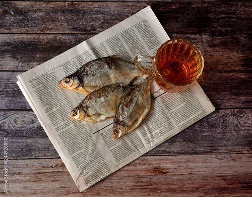 A glass mug of light beer, three dried river fish and an old newspaper on a dark wooden table. photo