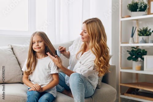 Mother gently combing daughter's hair on cozy living room couch, bonding moment in warm domestic setting