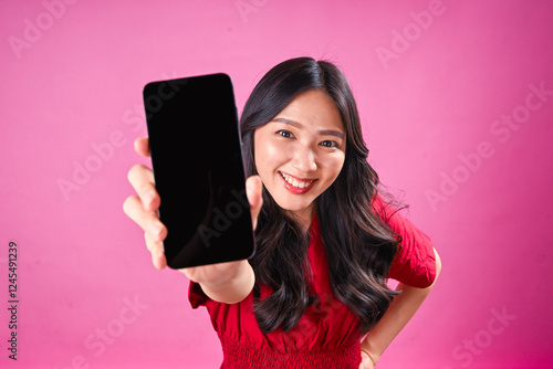 A cheerful young Asian woman with long wavy black hair wearing a red dress, smiling while extending a smartphone towards the camera, against a pink background