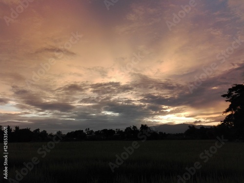 A breathtaking sunset over the countryside, with the sky painted in shades of orange, pink, and purple. The rice fields below are darkened by the fading light, while distant mountains create a majesti photo