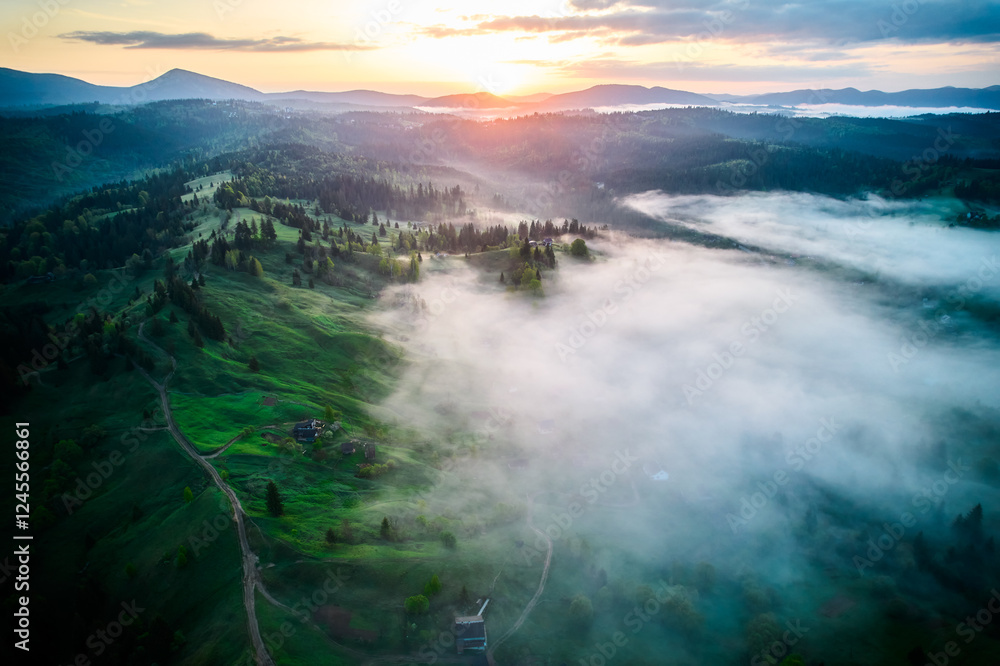 Obraz premium Aerial view of sunrise over mist-covered landscape, with golden light illuminating rolling hills and valleys. Thick fog creates mystical atmosphere, partially obscuring lush greenery and trees below.