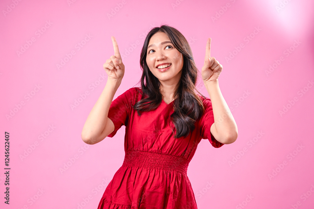 A young Asian woman with long wavy black hair wears a red dress and smiles brightly while pointing upwards with both hands against a pink background, expressing excitement A young Asian woman with long wavy black hair wears a red dress and smiles brightly while pointing upwards with both hands against a pink background, expressing excitement