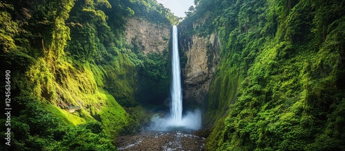 Fototapeta Naklejka Na Ścianę i Meble -  Majestic waterfall cascading down rocky cliffs, surrounded by lush green jungle foliage, under a clear blue sky with sunlight filtering through leaves.