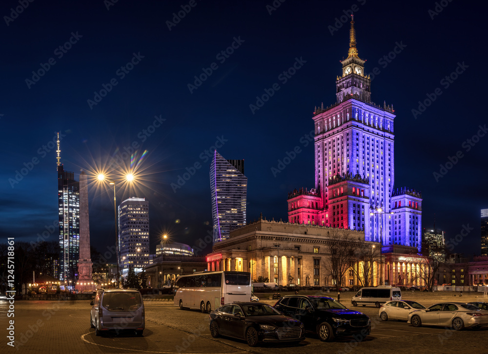 Naklejka premium Night in Warsaw, Poland. The Palace of Culture and Science is illuminated in vibrant red, blue, and purple lights, contrasting with the dark sky. City lights and modern skyscrapers complete scene.