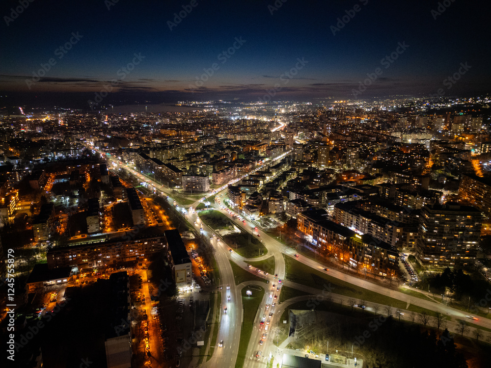 Obraz premium Aerial view of Varna, Bulgaria, at sunset, showcasing a vibrant cityscape with illuminated streets, moving traffic, and a glowing skyline. The coastal metropolis reflects urban energy and modern