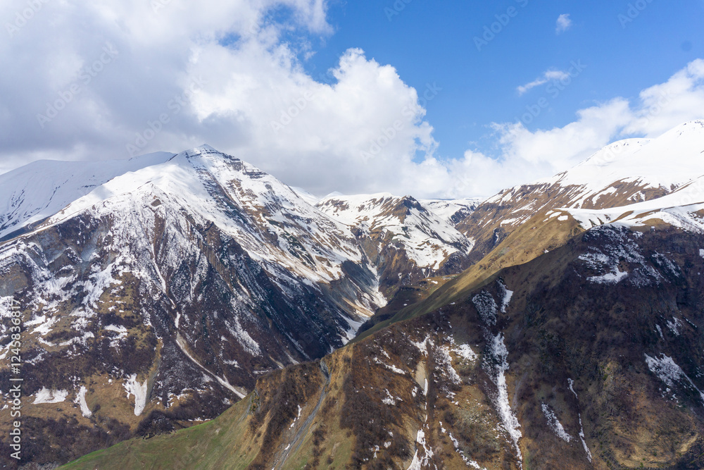 Fototapeta premium Snowy mountains covered with snow and green grass. Geological formations are visible on the rocks. Blue sky with clouds in the background