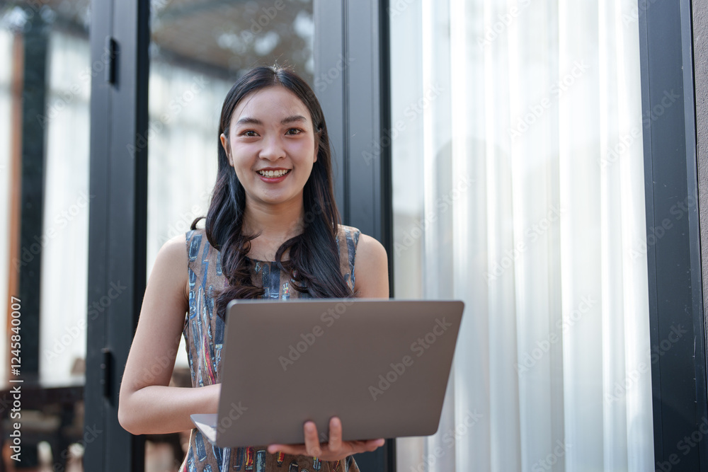 Young asian businesswoman smiling while using a laptop near a window in a modern office, embodying the essence of remote work and digital communication Young asian businesswoman smiling while using a laptop near a window in a modern office, embodying the essence of remote work and digital communication