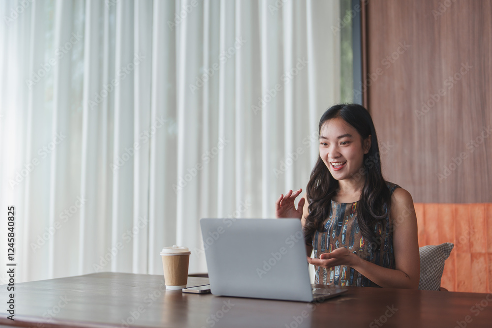 Happy young asian businesswoman having a video conference on laptop computer while sitting at her desk in modern office with a cup of coffee
