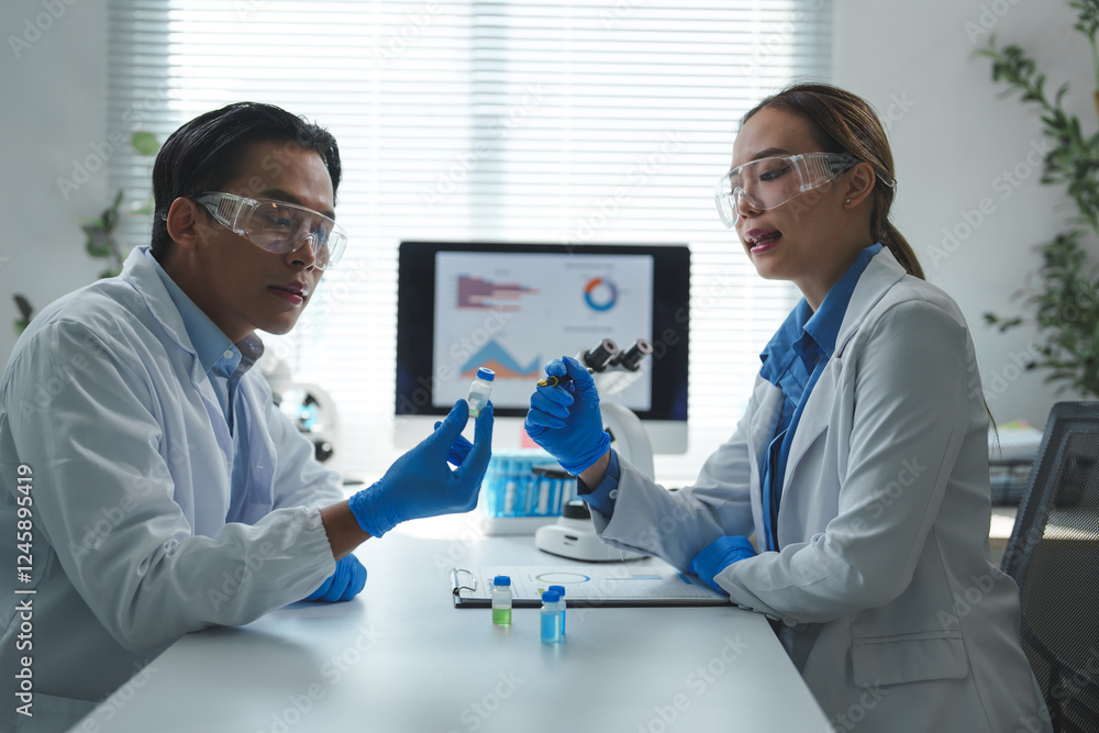 Two scientists in lab coats and safety glasses examining medical sample vials and discussing findings in a modern laboratory with a microscope and data analysis on a computer Two scientists in lab coats and safety glasses examining medical sample vials and discussing findings in a modern laboratory with a microscope and data analysis on a computer
