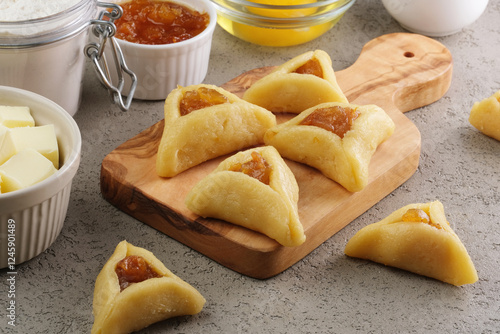 Raw sugar cookies in a triangular shape with filling on a wooden cutting board, preparation for the traditional Jewish holiday Purim day. A group of ingredients on the background. side view photo