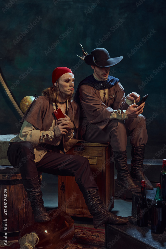 Fototapeta premium Young men, medieval pirates in vintage costumes sitting with smartphones, scrolling social media feed, discussing news against dark background in dimly-lit room. Concept of digital networking