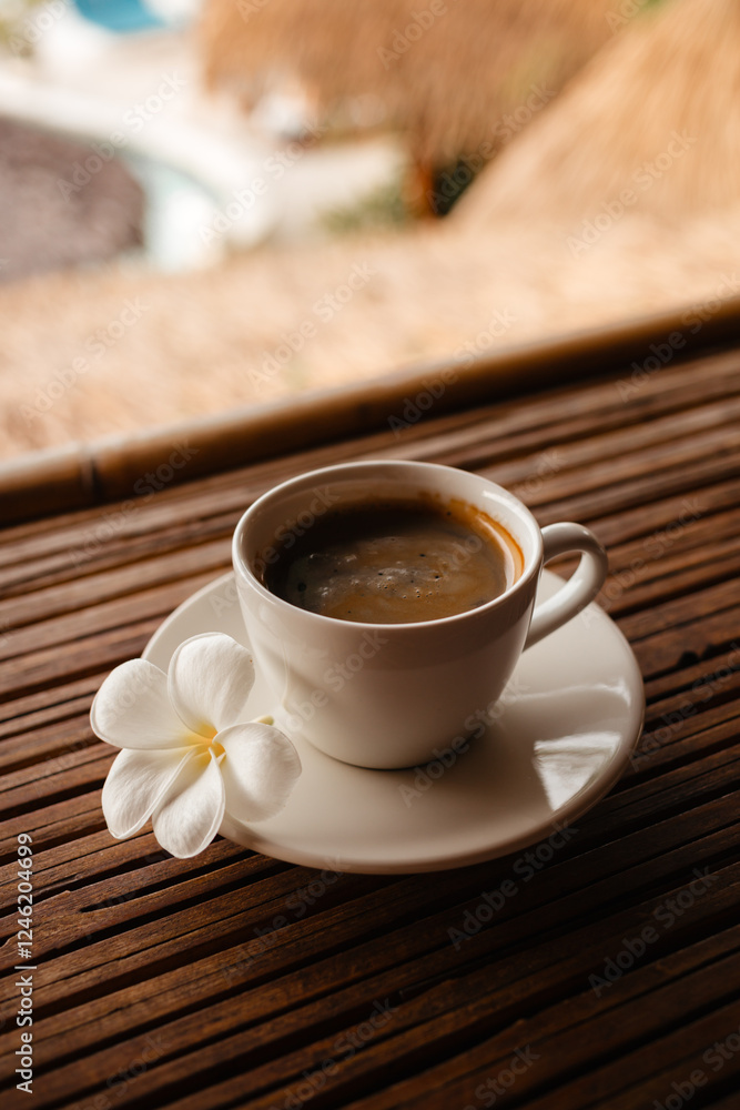 Morning hot Americano in a white mug with frangipani flower. Black coffee on a bamboo table. A cup of strong coffee in the morning on vacation Morning hot Americano in a white mug with frangipani flower. Black coffee on a bamboo table. A cup of strong coffee in the morning on vacation