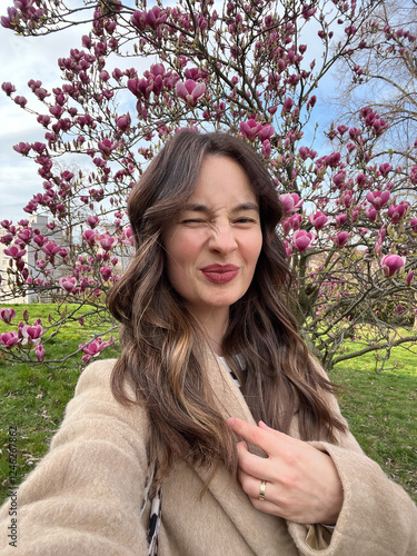 Caucasian woman with air touch hair coloring winks and taking a selfie near blooming magnolia tree photo