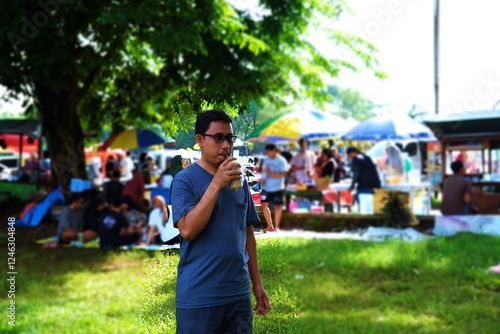 A man stands in a vibrant outdoor market, enjoying a refreshing drink on a sunny day. Surrounded by food stalls, greenery, and a lively crowd, this photo captures a relaxed moment in a festive setting photo