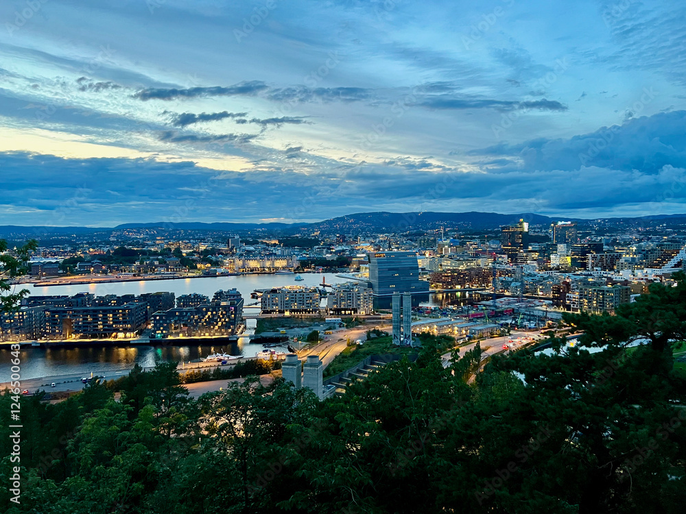 Fototapeta premium Evening panorama of Oslo city viewed from Ekebergrestauranten, showcasing illuminated cityscape, roads, hills, and dramatic twilight sky in Norway.