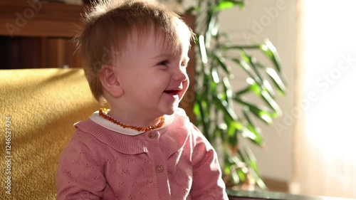 Adorable little baby girl smiles to camera while sitting on a yellow armchair in the living room. Home atmosphere, family, socialising, child, growing up, baby portrait.