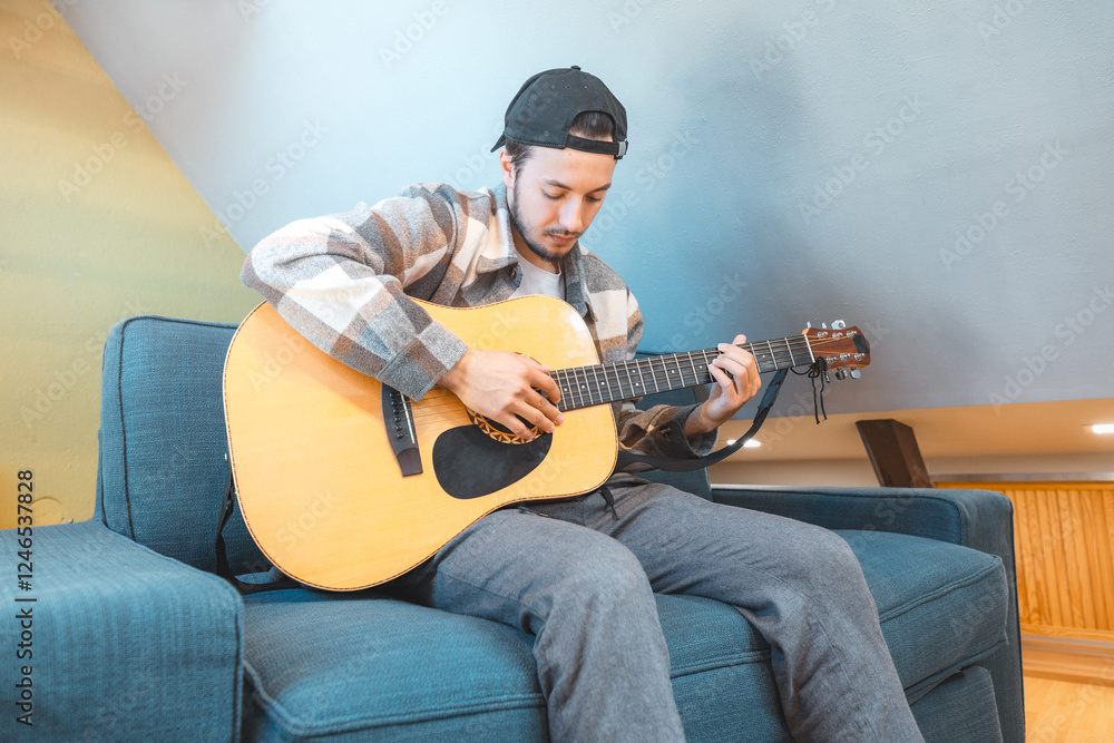Obraz premium Young man with a relaxed demeanor sits barefoot on a cozy blue couch, strumming an acoustic guitar. His expression shows contentment and focus as he connects with his music. Casual and creative vibe