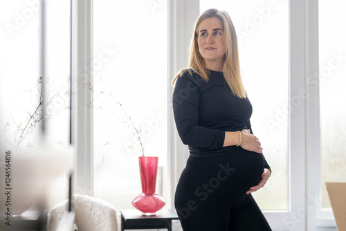 A pregnant woman in a black outfit stands in a bright room, gently cradling her belly. Netherlands photo