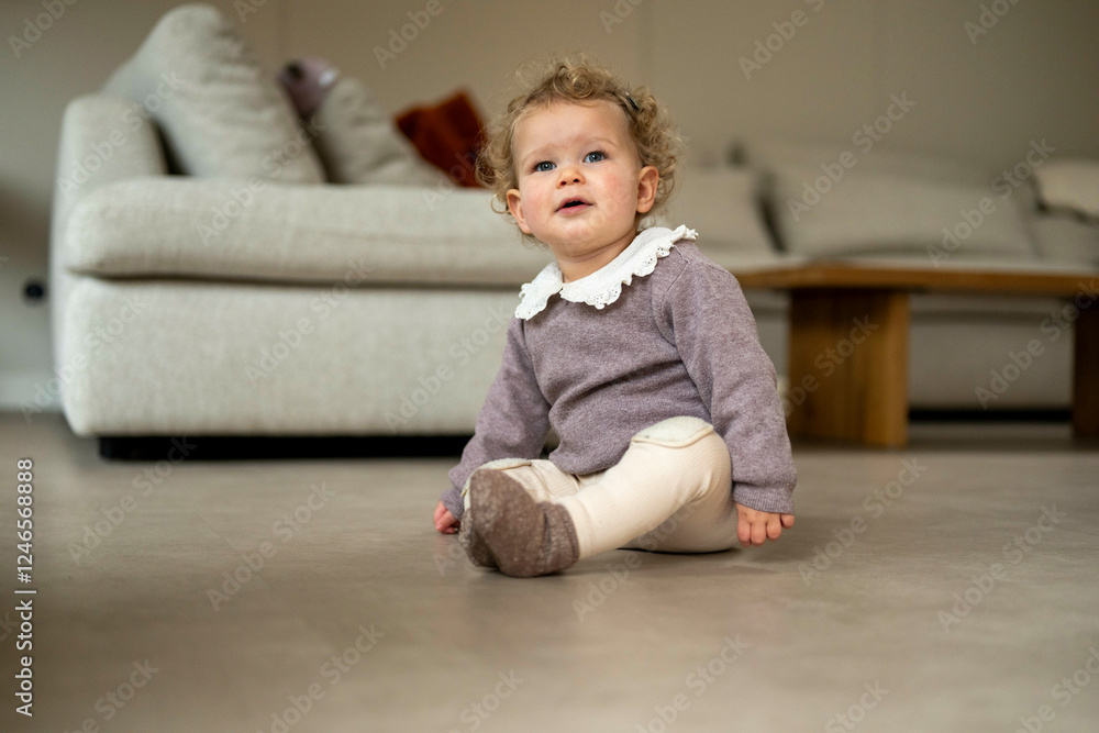 Curly-haired toddler in cozy clothes sits on floor with beige sofa in background. Netherlands Curly-haired toddler in cozy clothes sits on floor with beige sofa in background. Netherlands