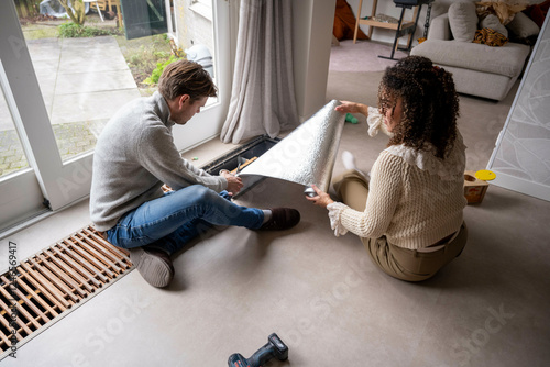 Man kneels installing flooring and isolation while woman assists in a bright, modern living room. Netherlands