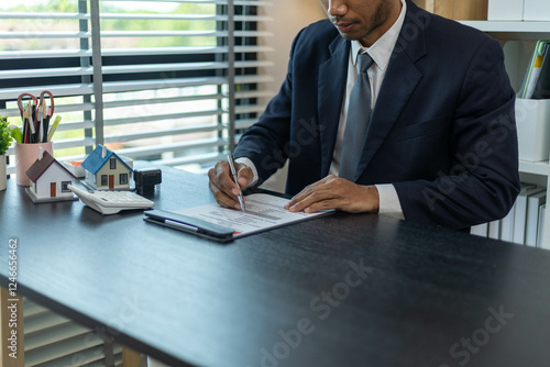 A man is sitting at a desk with a calculator and a piece of paper with numbers on it. He is pointing to the paper and he is explaining something to someone. The scene suggests a professional setting

