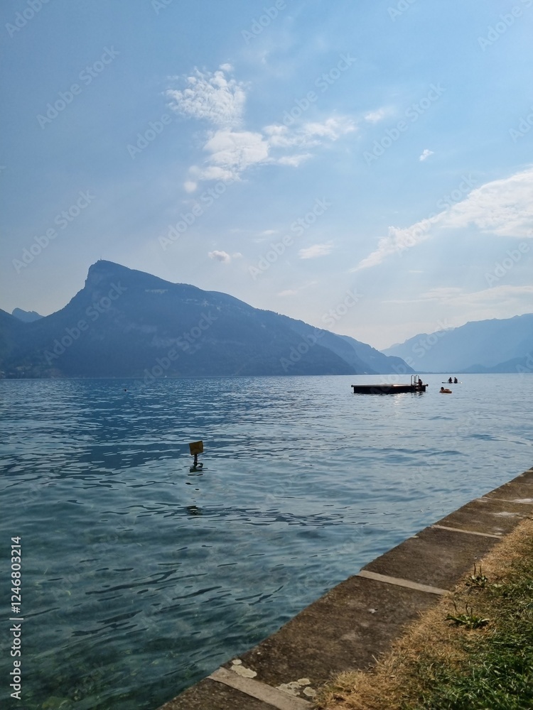 Obraz premium Serene lake landscape with a boat anchored in the calm water, a majestic mountain range in the background, clear blue sky with scattered clouds, and a stone pier stretching into the lake.