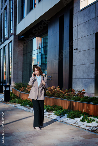 A cheerful young businesswoman in a stylish blazer is having a phone conversation outdoors in an urban setting. She is smiling confidently, exuding positivity and professionalism. 