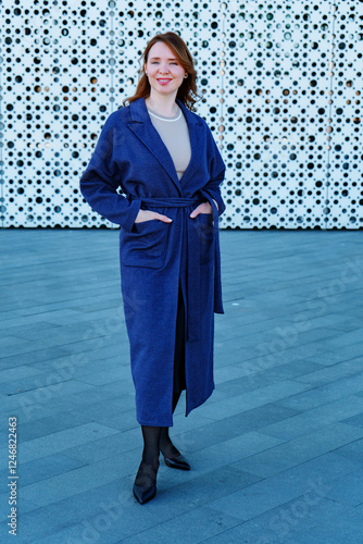 Elegant woman in a stylish blue coat standing against a modern architectural background. She smiles confidently, exuding warmth and sophistication. Outdoor portrait with natural lighting.