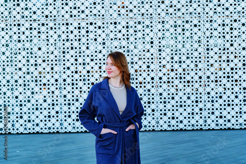 Elegant woman in a stylish blue coat standing against a modern architectural background. She smiles confidently, exuding warmth and sophistication. Outdoor portrait with natural lighting.