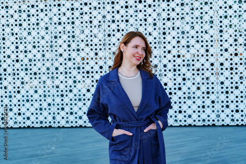 Elegant woman in a stylish blue coat standing against a modern architectural background. She smiles confidently, exuding warmth and sophistication. Outdoor portrait with natural lighting.