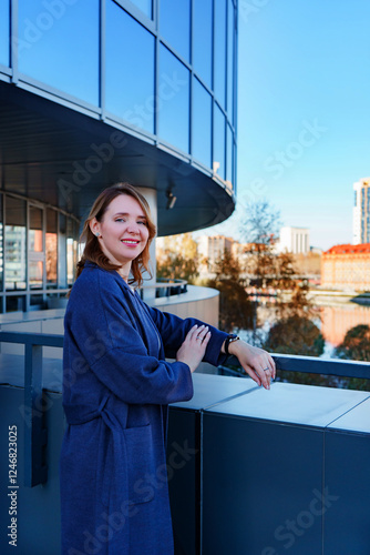 A stylish young woman in a blue coat stands outside a modern glass building, leaning on a railing and smiling. The reflective windows of the structure capture the surrounding cityscape.