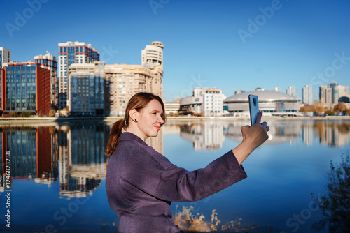 Young woman taking a selfie with a smartphone against a modern cityscape and water reflections. Concept of urban lifestyle, tourism, social media, and digital connection.