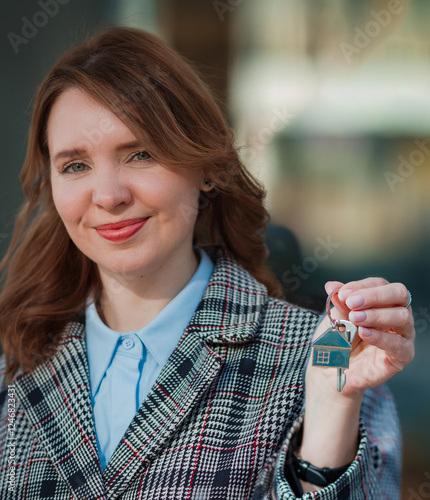 Smiling woman holding house keys with a home-shaped keychain. Represents real estate, home purchase, renting, or investment. Modern city background, professional and confident look.