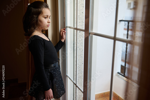 Young girl in black dress standing by a window and looking outside thoughtfully photo