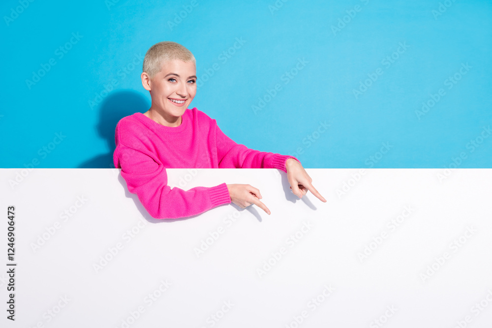 Young woman with a trendy short blonde hairstyle in a bright pink sweater pointing downwards against a vibrant blue background Young woman with a trendy short blonde hairstyle in a bright pink sweater pointing downwards against a vibrant blue background