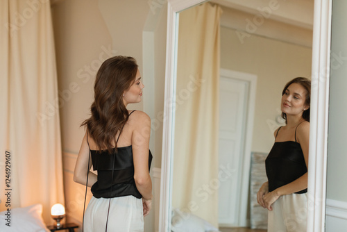 Stylish young woman standing in front of a mirror in her bedroom looking at her reflection