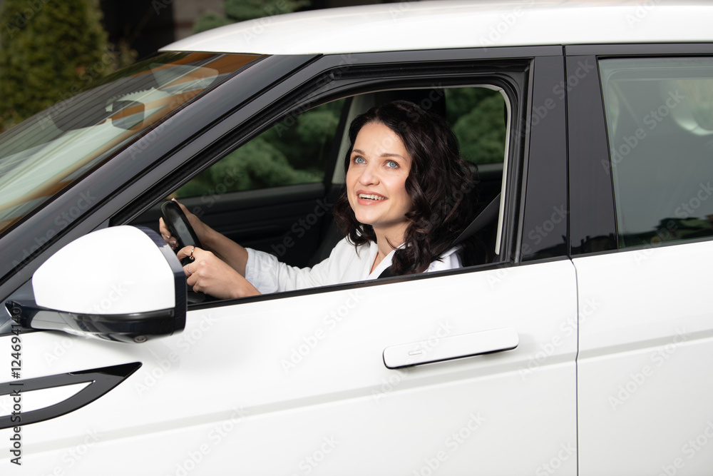 A cheerful woman with curly hair is seen behind the wheel of a white car parked in a neighborhood. Sunlight shines through the window as she smiles confidently A cheerful woman with curly hair is seen behind the wheel of a white car parked in a neighborhood. Sunlight shines through the window as she smiles confidently