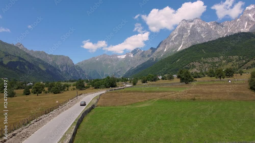 Gray station wagon drives along winding dirt road through green meadows surrounded by wooded mountains under blue sky with some clouds. shooting from drone. Gray station wagon drives along winding dirt road through green meadows surrounded by wooded mountains under blue sky with some clouds. shooting from drone.