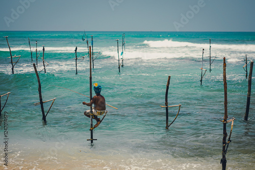 Rear view of a fisherman fishing on a stilt in the sea in Sri Lanka. A young man wearing an blue turban sits on a wooden pole that is unique to this country on a sunny day. Idea for unseen travel.