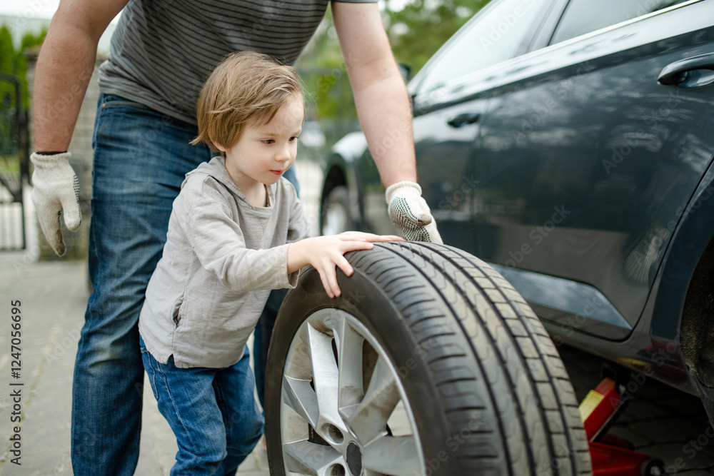 Cute little boy helping his father to change car wheels at their backyard. Father teaching his little son to use tools. Cute little boy helping his father to change car wheels at their backyard. Father teaching his little son to use tools.
