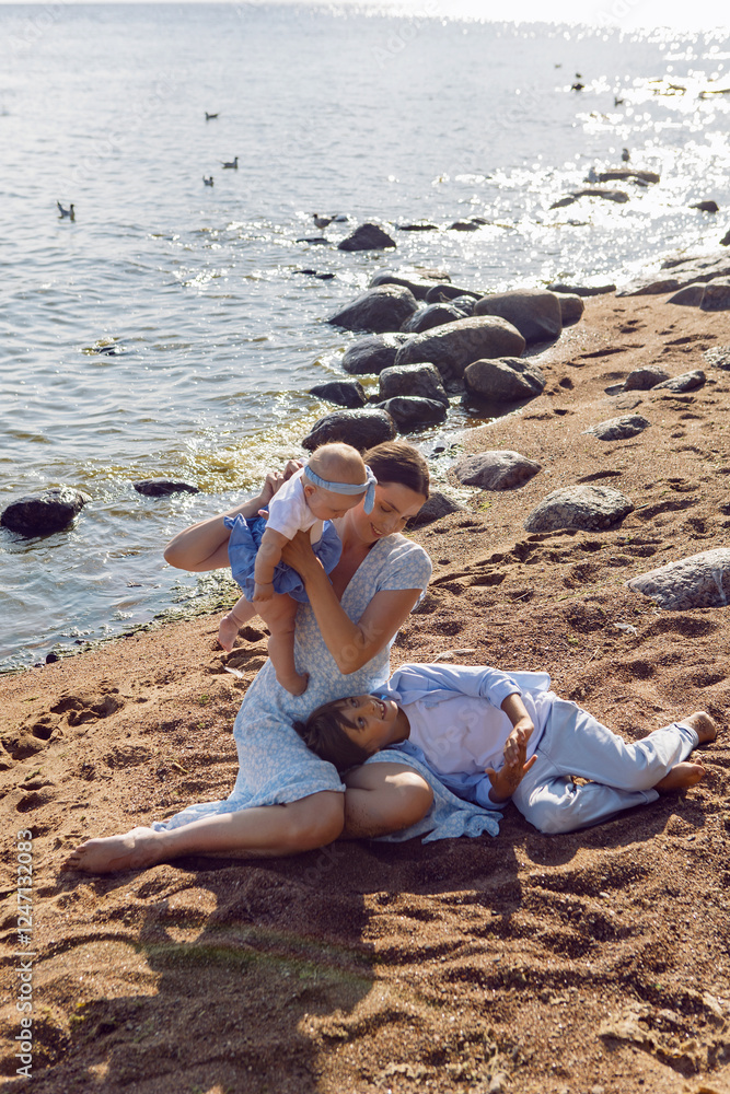 A happy mother in a dress with two children aged seven and a baby are standing on a sandy beach by the sea in summer
