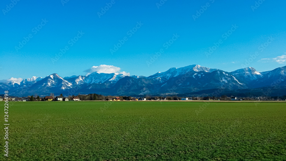 Obraz premium Green meadow stretches towards small village of St. Egyden in Carinthia, Austria. Majestic snow-capped Karawanks mountain range creating dramatic backdrop under pristine clear blue sky. Austrian Alps