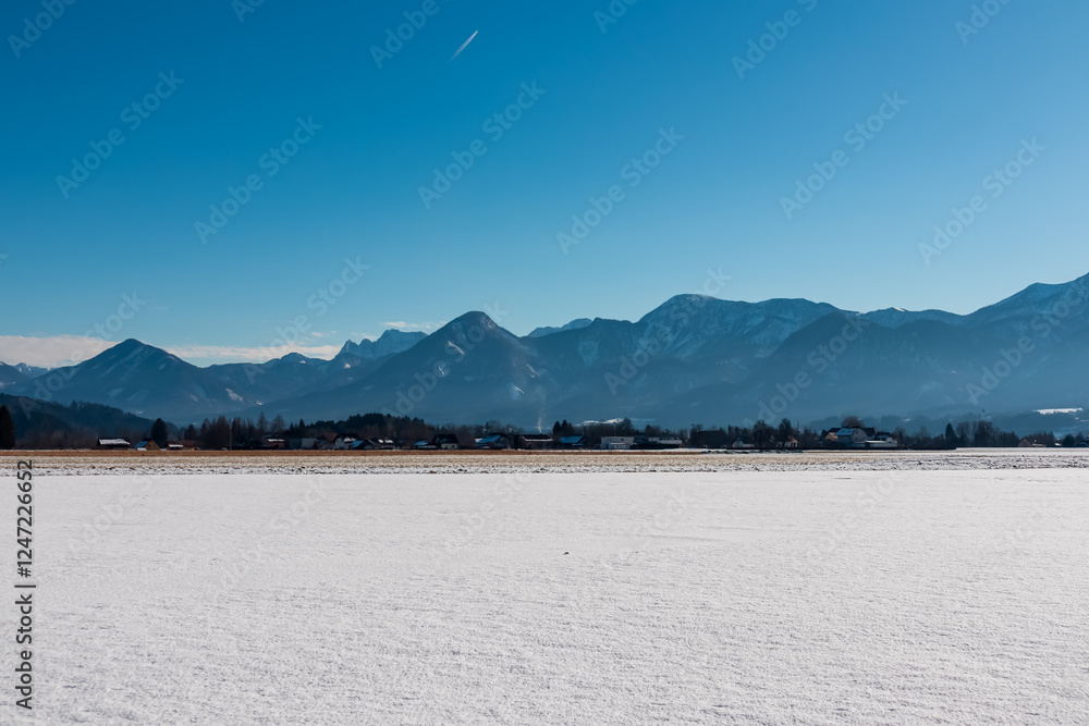 Obraz premium Serene winter landscape in St. Egyden, Carinthia, Austria. Snow-covered fields and majestic Karawanks mountain range rising in background. Small town in valley under clear blue sky. Wild Austrian Alps